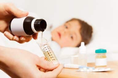 Un parent verse un médicament liquide dans un doseur, tandis qu’un jeune enfant malade est allongé en arrière-plan.