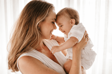 Maman souriante tenant son nourrisson dans ses bras, leurs fronts collés l’un contre l’autre, dans une lumière douce et chaleureuse évoquant un moment de tendresse et de complicité.