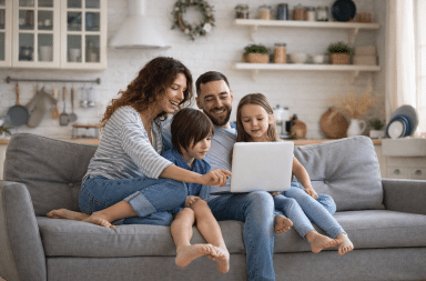 Famille de quatre personnes assise sur un canapé, les parents et leurs deux enfants regardant un ordinateur portable ensemble dans un salon lumineux, illustrant les démarches en ligne pour les allocations familiales.